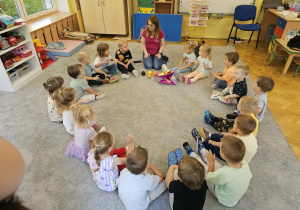 Dzieci siedzą na dywanie w kole i uczestniczą w zajęciach języka angielskiego.