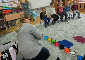 Pani Beata zaprasza dzieci do zabawy. Na dywanie w jednym rzędzie ustawione są miseczki Lili.
