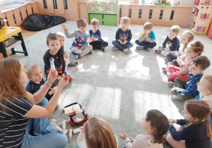 Grupa dzieci siedząc na dywanie ogląda skład apteczki pokazywanej przez nauczycielkę.