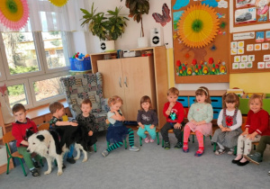 Lili przechodzi przed dziećmi siedzącymi na krzesełkach ustawionymi w półkolu. Dzieci delikatnie głaszczą psa.