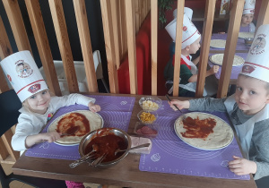 Dzieci siedząc przy stole smarują pizze sosem.
