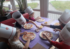 Dzieci siedząc przy stole dekorują pizze różnymi składniami.