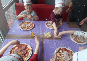 Dzieci siedząc przy stole dekorują pizze składnikami.