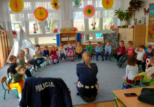 Dzieci siedzą na krzesełkach ustawionych na dywanie w półkolu. Gość siada przed dziećmi- spotkanie rozpoczyna zagadką.