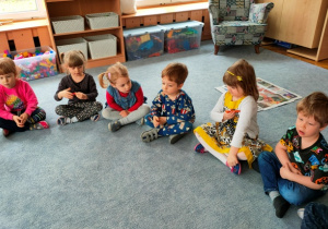Dzieci siedzą w półkolu na dywanie . Dziewczynka pokazuje dzieciom, jak mierzy się temperaturę.