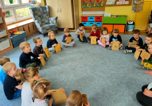 Dzieci siedzą w kole na dywanie. W rękach trzymają prezenty - koperty w których znalazły się przybory wykorzystywane przez nie w zabawach.
