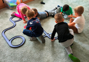 Dzieci ułożyły na dywanie tor dla małych samochodów. Bawią się zgodnie w zespole rówieśniczym.