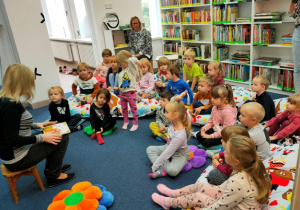 Dzieci siedzą na kolorowych poduchach ułożonych na dywanie- słuchają bajki o Jesieni czytanej przez panią bibliotekarkę.