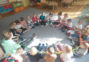Dzieci siedzą na dywanie tworząc koło z pomocą gumy sensorycznej.