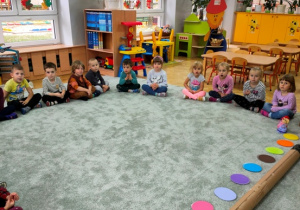Dzieci siedzą na dywanie w półkolu. Przed nimi położona jest duża tekturowa tuba, pod którą ułożone są krążki w 10 kolorach.