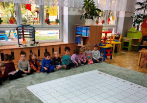 Dzieci siedzą na dywanie, patrzą na rozłożoną przed nimi matę do kodowania. Słuchają wyjaśnień nauczycielki.