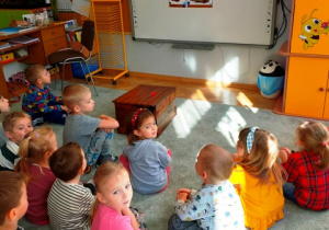 Dzieci siedzą na dywanie w luźnej gromadce. Słuchając opowiadania cioci o zwierzętach , oglądają ilustracje umieszczone na tablicy.
