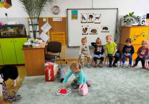 Dziewczynka nalewa wodę do psiej miski, pozostałe dzieci siedzą w półkolu na krzesełkach ustawionych na dywanie.