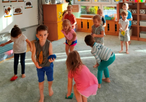 Dzieci stoją w miejscu na dywanie - podrzucają woreczek i próbują go złapać.