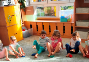 Dzieci siedzą w siadzie skulnym. Palcami stóp próbują podnieść woreczek z dywanu.