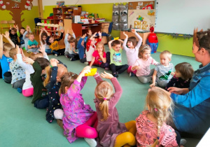 Dzieci siedzą w dwóch rzędach, ręce unoszą nad głowy. Przekazują nad głowani woreczek jeżowych przysmaków.
