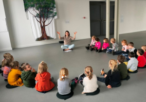 Dzieci siedzą w dużym półkolu. Słuchają wypowiedzi prelegenta o prezentowanym dziele sztuki- wyhaftowanym drzewie.
