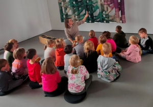 Dzieci siedzą w luźnej gromadce. Rozmawiają z prowadzącym o oglądanym dziele sztuki współczesnej.