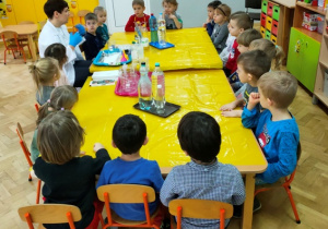 Dzieci siedzą przy połączonych stołach. Słuchają gościa, który opowiada na czym polega jego praca- praca chemika.
