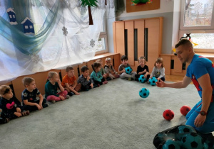 Dzieci siedzą na dywanie w jednym rzędzie. Trener kolejno podaje dzieciom piłki- poznaje imiona uczestników zabaw.