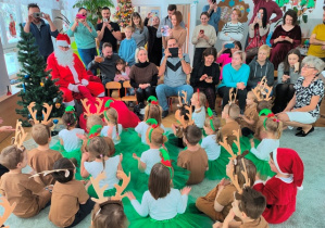 Dzieci siedzą na dywanie zwrócone twarzami do Mikołaja, śpiewają mu piosenkę.