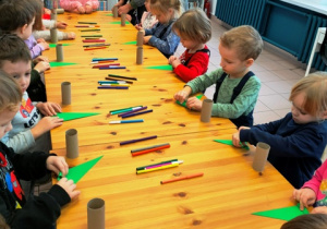 Dzieci stoją przy stolikach na których przygotowane są materiały plastyczne- składają zielony trójkąt w harminijkę.