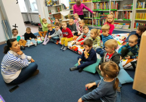 Dzieci siedzą na kolorowych poduchach w czytelni. Rozmawiają z panią bibliotekarką o ulubionych zimowych zabawach.