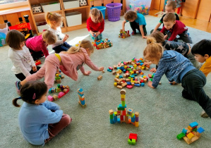 Dzieci skupione na dywanie. Budują z drewnianych klocków wymyślone przez siebie budowle.