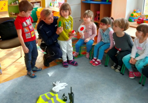 Dzieci stoją obok policjantki, słuchają zadania przygotowanego przez gościa.