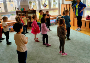 Dzieci stoją w rozsypce na dywanie- rozgrzewają ciało naśladując instruktora tanca.