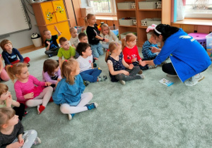 Dzieci siedzą w siadzie skrzyżnym na dywanie. Instruktorka żegna się kolejno z każdym dzieckiem umieszczając na jego dłoni okolicznościowy stempelek.