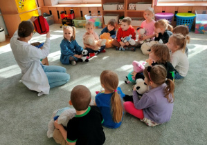 Grupa dzieci wraz ze swoimi przytulankami siedzi w półkolu na dywanie- słucha wypowiedzi gościa.