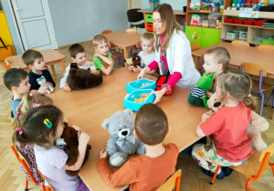 Grupa dzieci siedzi przy stoliku- studentka medycyny prezentuje dzieciom akcesoria lekarskie.