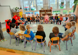 Dzieci siedzą na krzesełkach w dużym kole. Słuchają wypowiedzi gościa.