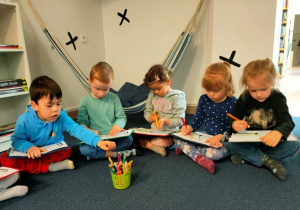 Dzieci siedzą na dywanie. Na kartce- książce umieszczonej na podkładce tworzą ilustrację do wymyślonej przez siebie opowieści.