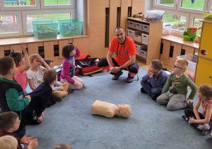 Dzieci siedzą na dywanie i słuchają o pracy ratownika medycznego.