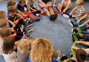 Dzieci siedząc na dywanie trzymają gumę sensoryczną w dłoniach i poruszają nią od stóp do kolan