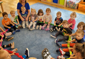 Dzieci siedzą na dywanie tworząc koło przy pomocy gumy sensorycznej