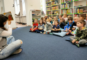 Dzieci siedząc na poduchach w czytelni, słuchają bajki czytanej przez panią Olę.