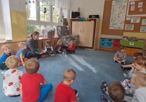 Dzieci siedzą na dywanie i uważnie słuchają opowiadania czytanego przez ciocię.