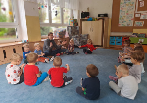 Dzieci siedzą na dywanie i razem z ciocią śpiewają piosenkę.