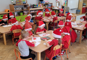 Dzieci siedzą przy stolikach i malują czerwoną farbą mikołajkowe czapeczki.