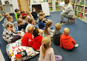 Dzieci siedzą na poduchach w czytelni. Słuchają bajki czytanej przez panią bibliotekarkę.