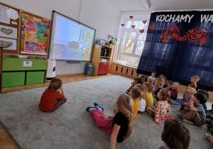 Dzieci siedzą na dywanie i oglądają prezentację