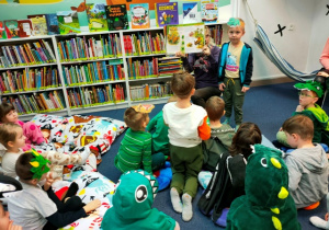 Dzieci wspólnie z panią bibliotekarką szukają rodziny dinozaura znajdującego się na emblemacie chłopca.