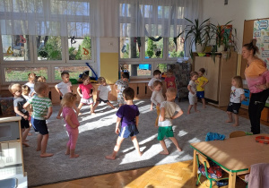 Dzieci stoją na dywanie w rozsypce i rozgrzewają biodra kręcąc nimi w prawo i w lewo.