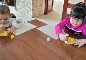 Dziewczynki przyklejają na karton żółte jajeczka wycięte z kolorowego papieru.