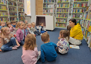 Dzieci siedzą grzecznie na dywanie i słuchają zasad w bibliotece.