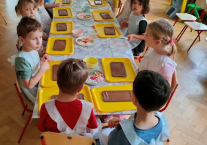 Dzieci siedzą przy stolikach, przed nimi leżą formy z masą czekoladową.