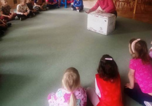 Dzieci siedzą na dywanie w dużym kole. Ciocia rozpakowuje pudło-niespodziankę.